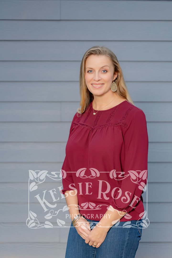 Smiling blonde woman in burgundy blouse standing against gray siding wall, professional portrait photography.