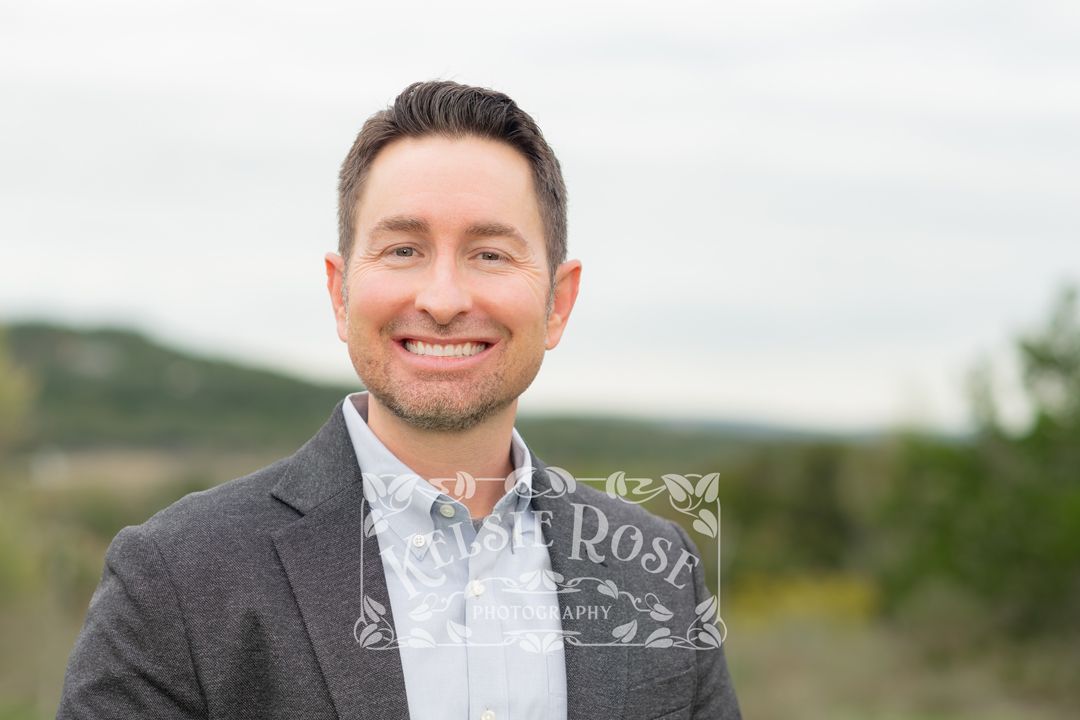 Headshot of professional man in gray blazer smiling outdoors with Texas hill country and cloudy sky in background.