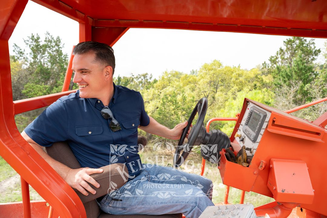 Smiling man in navy shirt operating controls of large orange road paving machine.