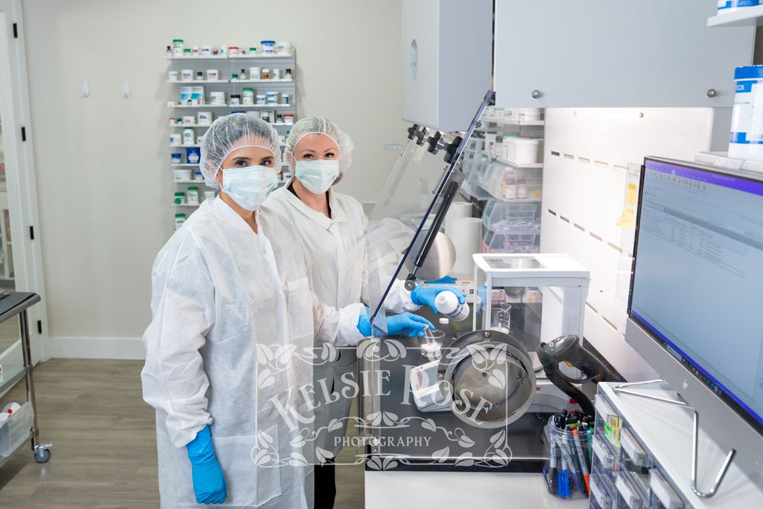 Two pharmacy technicians in white coats, masks, and gloves working with compounding equipment in clean lab.
