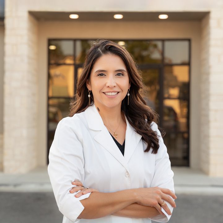Professional woman in white lab coat smiling with arms crossed outside a modern building entrance.