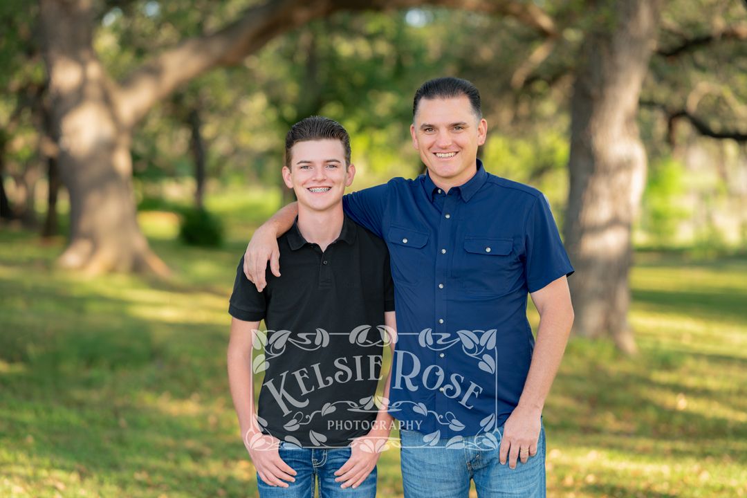 Father and son posing together outdoors with green oak trees in the background.