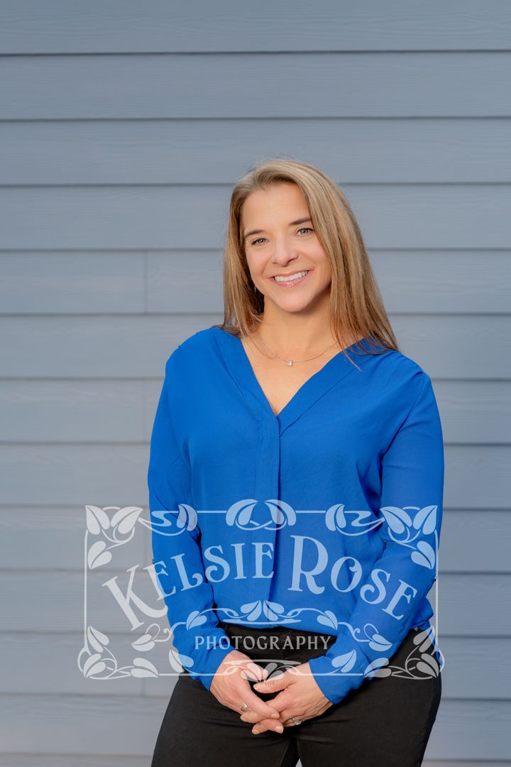 Smiling woman in royal blue blouse posing against gray siding wall, professional portrait photography.
