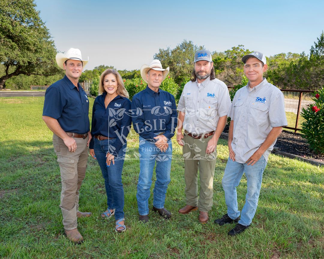 Group of five employees in uniform pose outdoors with grass and oak trees in the background in a rural Texas setting.