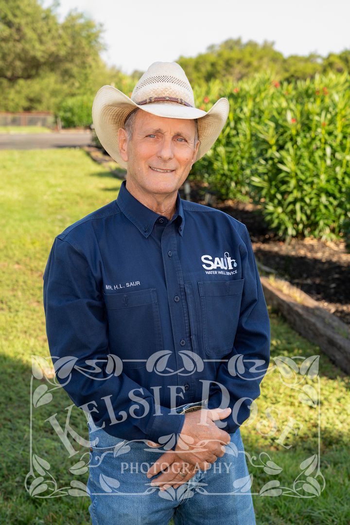 Man in cowboy hat and navy shirt smiles outdoors near oleander bushesin a rural Texas setting.