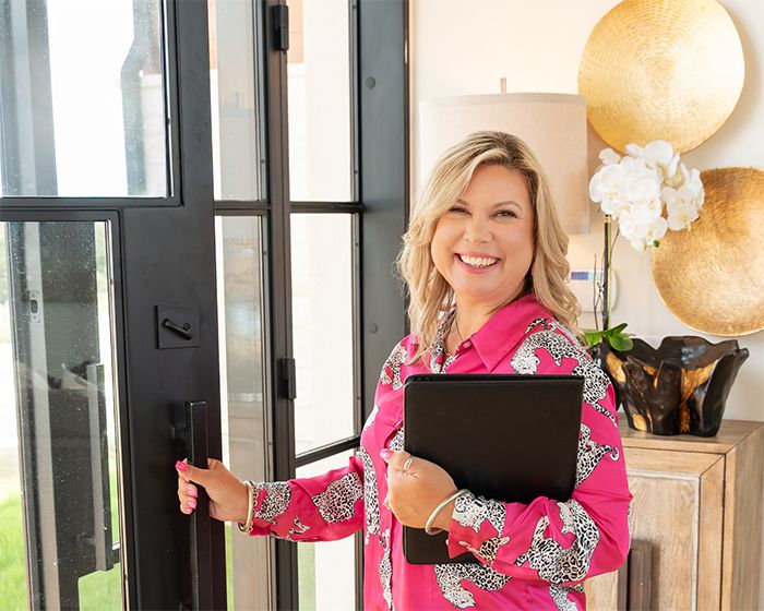 A smiling blonde woman in a pink floral blouse holds a leather folder while opening a modern black door.