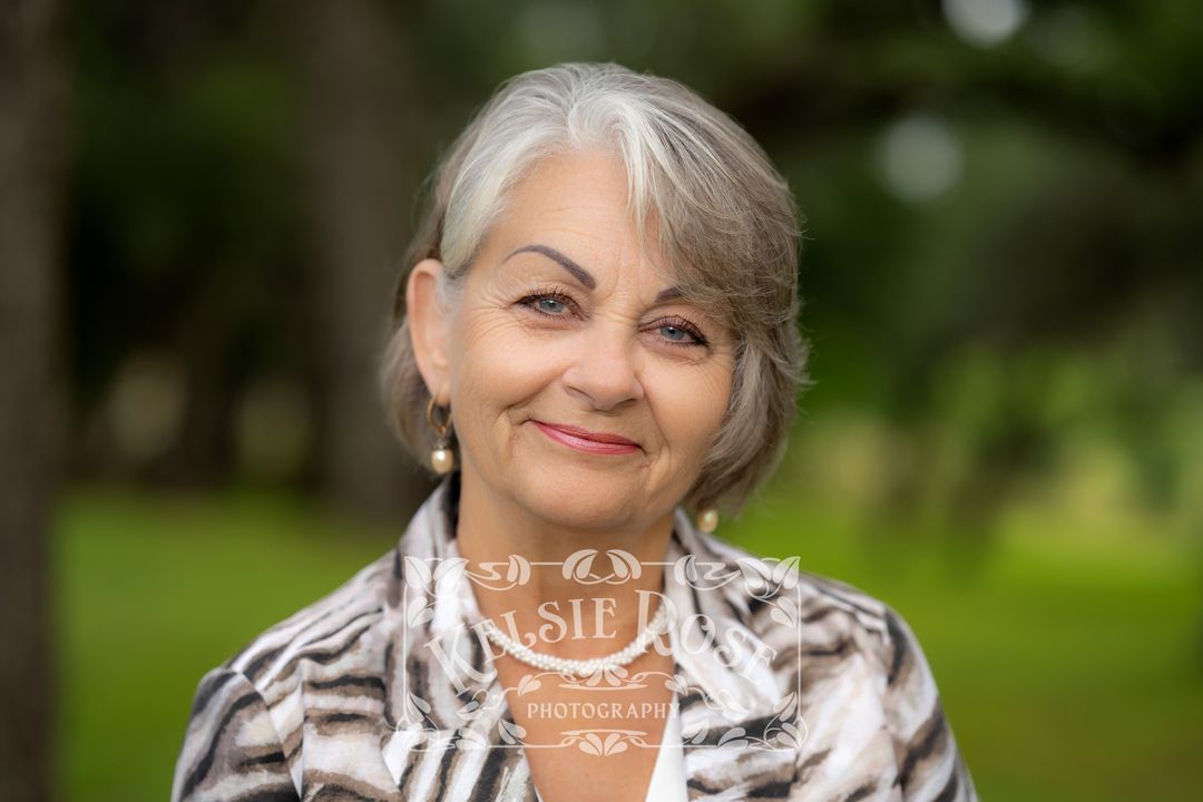 Headshot of smiling older woman with short gray hair wearing a patterned jacket outdoors with green oak trees in background.