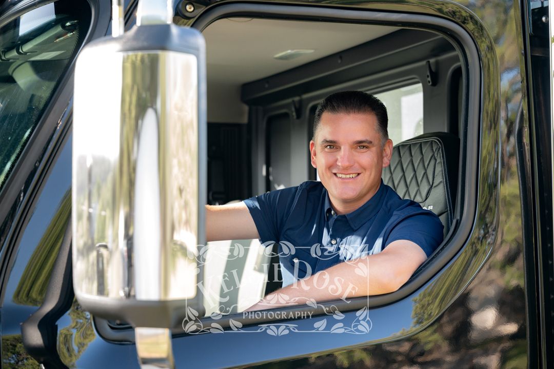 Professional portrait of smiling man in navy shirt leaning out of a large commercial truck cab window.