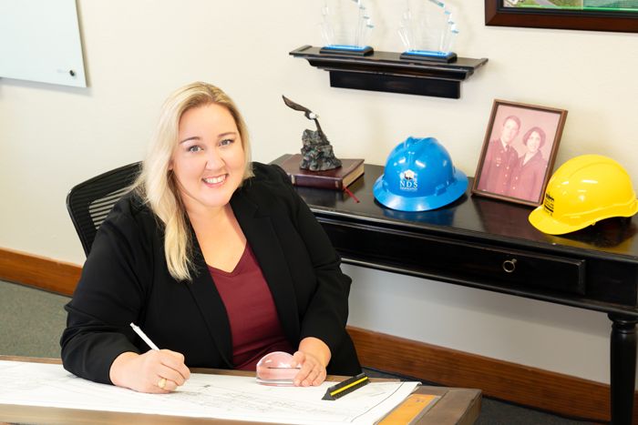 Professional woman in a black blazer sitting at a drafting desk with construction hard hats in an office.