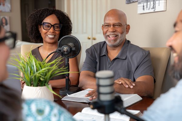 Two smiling adults recording a podcast episode together at a table with microphones and open Bible with second couple.