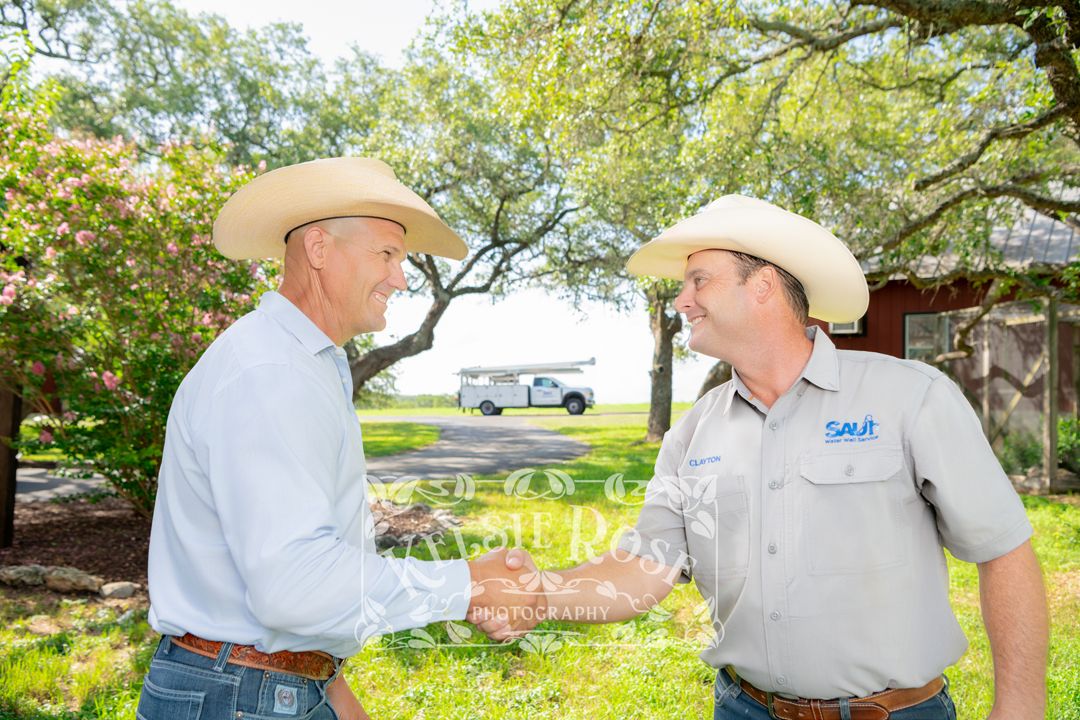Two men in cowboy hats shaking hands warmly in a green outdoor setting on a ranch in Texas.