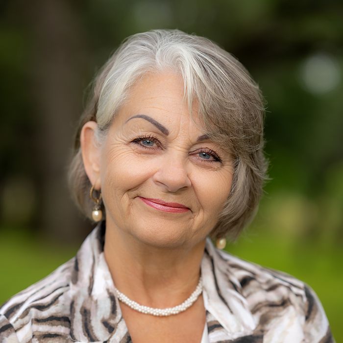 Smiling older woman with short gray hair wearing a pearl necklace and patterned blazer outdoors.