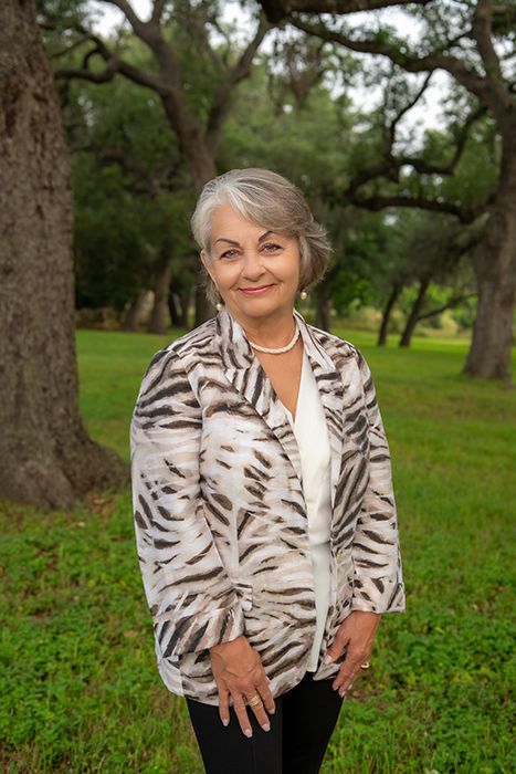 Elegant older woman with silver hair wearing a tiger-print blazer standing among oak trees.