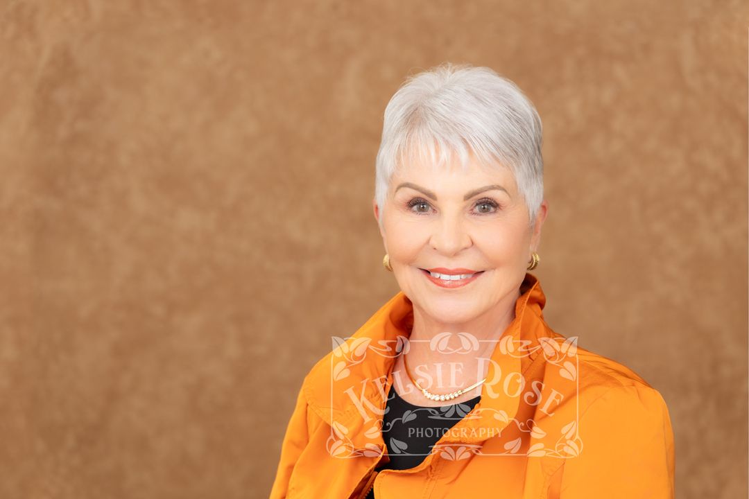Smiling senior woman with short white hair wearing an orange jacket, posing for a professional portrait photo.