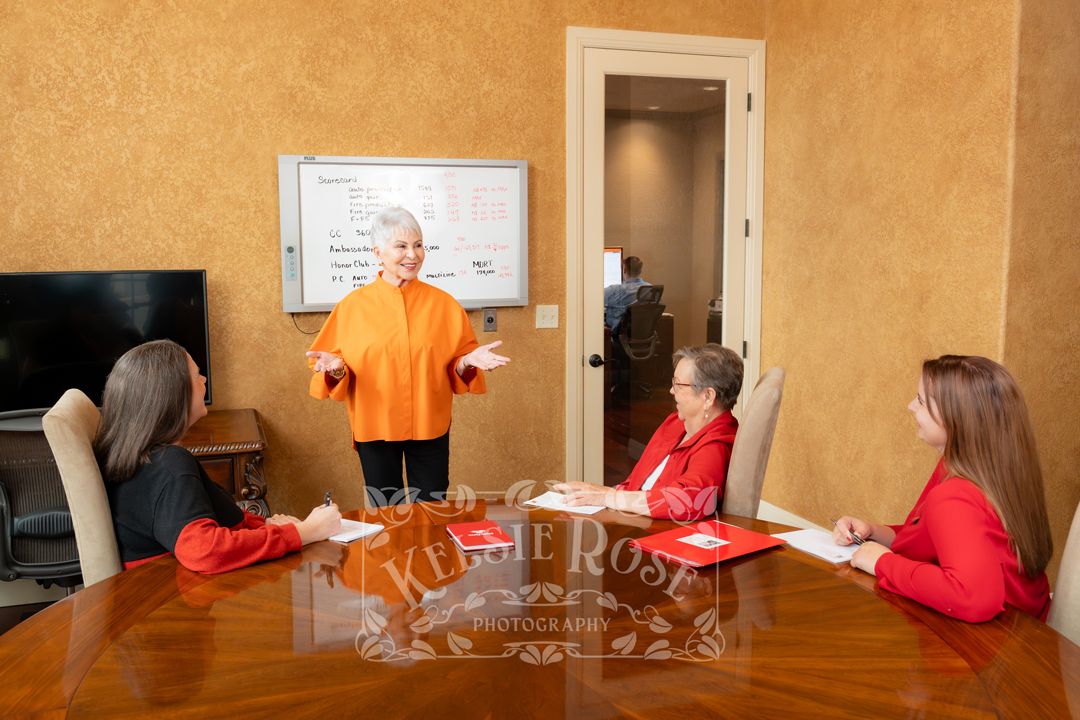 Woman in orange presents at whiteboard to three seated colleagues in a professional conference room meeting.