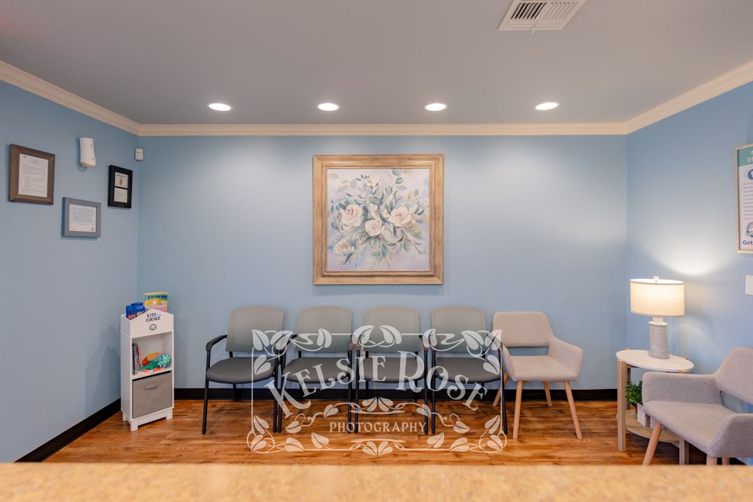 A bright dental office waiting room with blue walls, gray chairs, hardwood floors, and floral artwork.