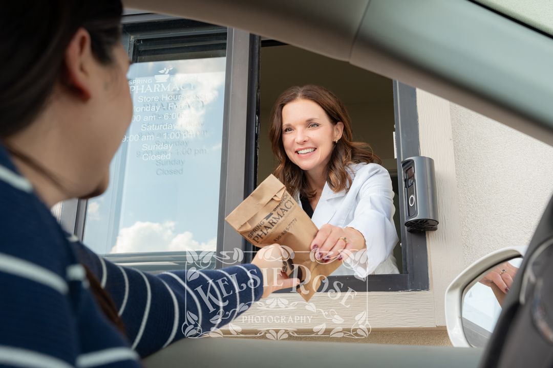 Smiling pharmacist in white coat handing a prescription bag to a customer at a drive-through window.