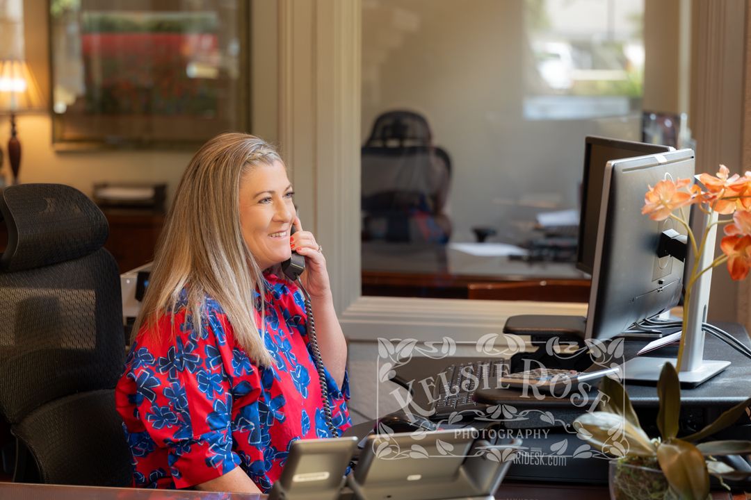Woman in red floral blouse smiling while talking on phone at elegant office desk with computer monitor.