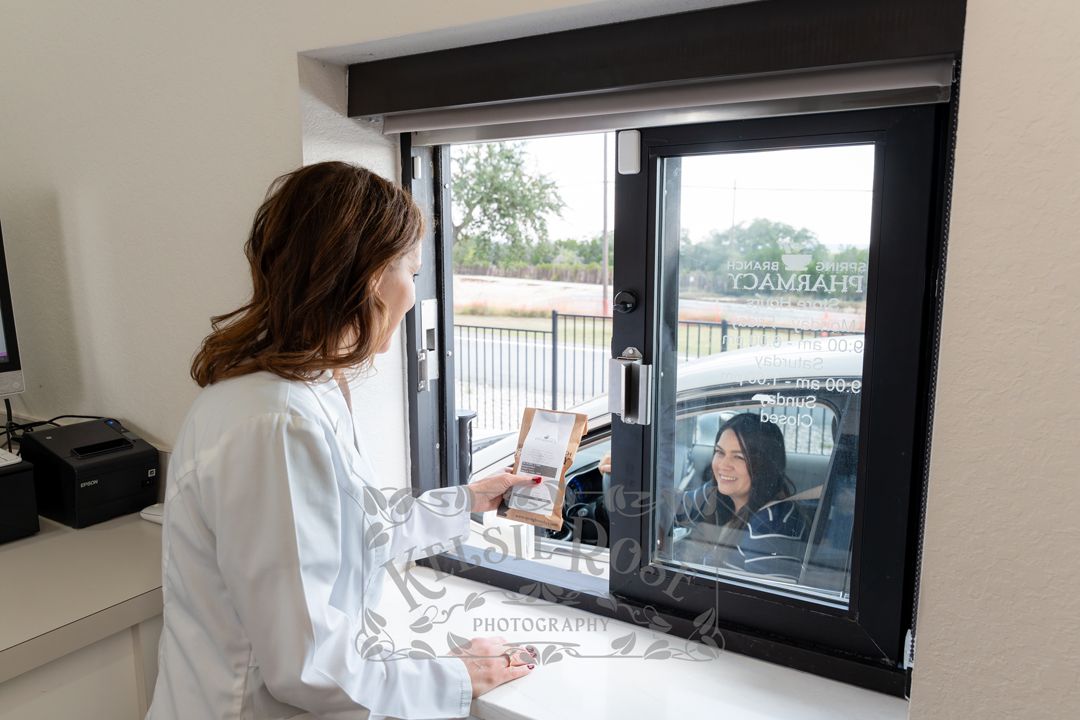 Female pharmacist in white coat assisting a drive-through pharmacy customer at a service window.