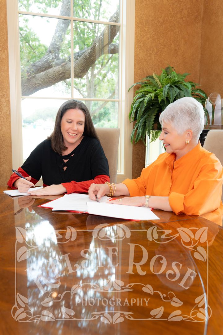 Two women smiling and signing documents at a polished wooden table near a bright window.