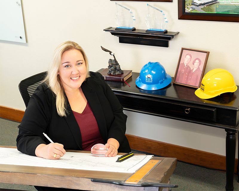 Professional woman smiling at drafting desk with construction hard hats and blueprints in office.