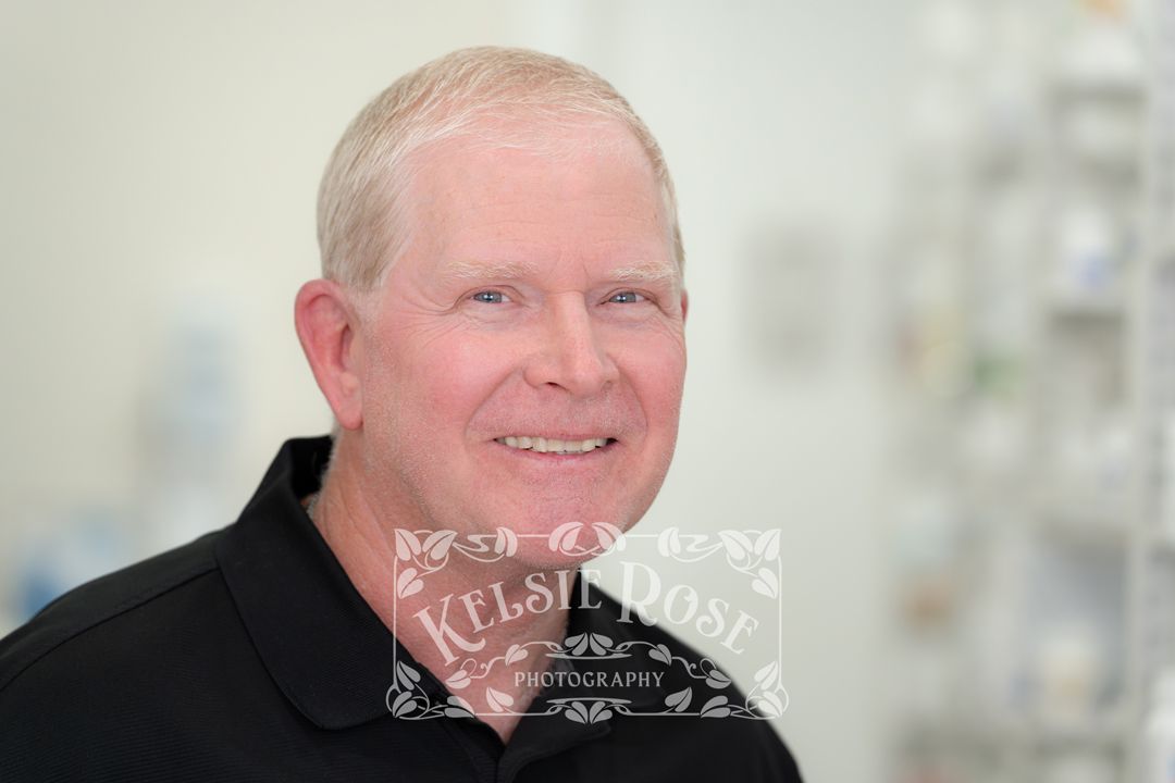 Headshot of a smiling male pharmacy employee in a black polo shirt inside a pharmacy.