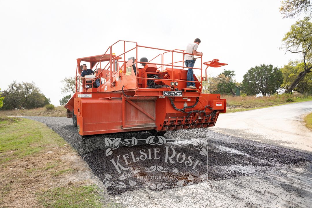 Workers operating large orange paving machine spreading asphalt on a rural road.