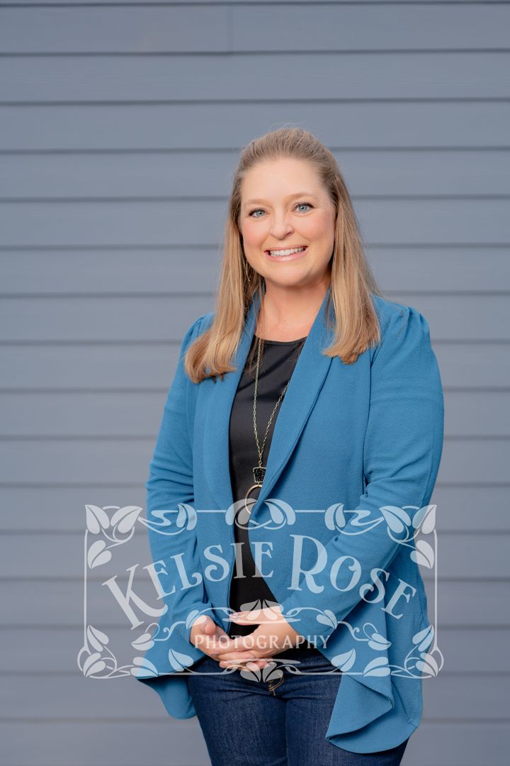 Smiling blonde woman in teal blazer and black top posing against gray siding, professional portrait photography.