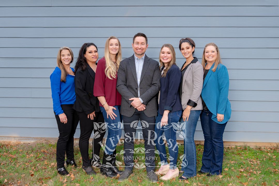 Group of seven professionals smiling together outdoors against a gray wall, team portrait photo.