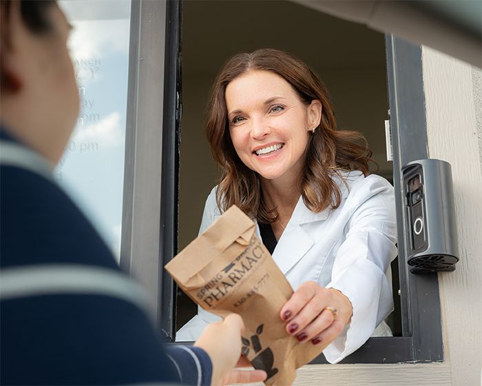 Smiling woman in white coat handing a brown paper bag through a drive-thru window to a customer.