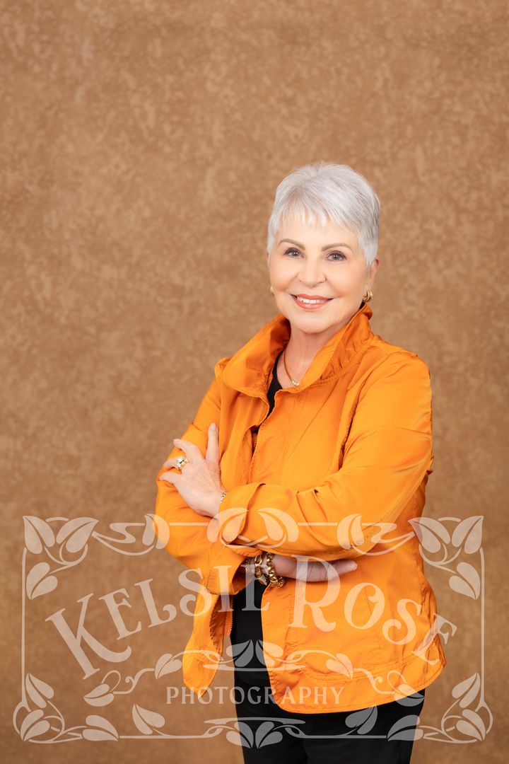 Smiling older woman with silver hair in orange jacket, arms crossed, posed against brown backdrop in professional portrait.