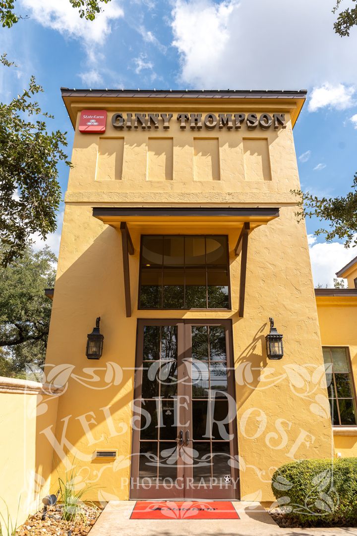 Yellow stucco building entrance with brown double doors under a canopy, featuring a Ginny Thompson sign and wall lanterns.