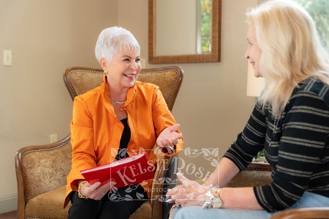 Two women smiling and conversing, one in an orange jacket holding a red book, seated in a cozy, well-lit room.
