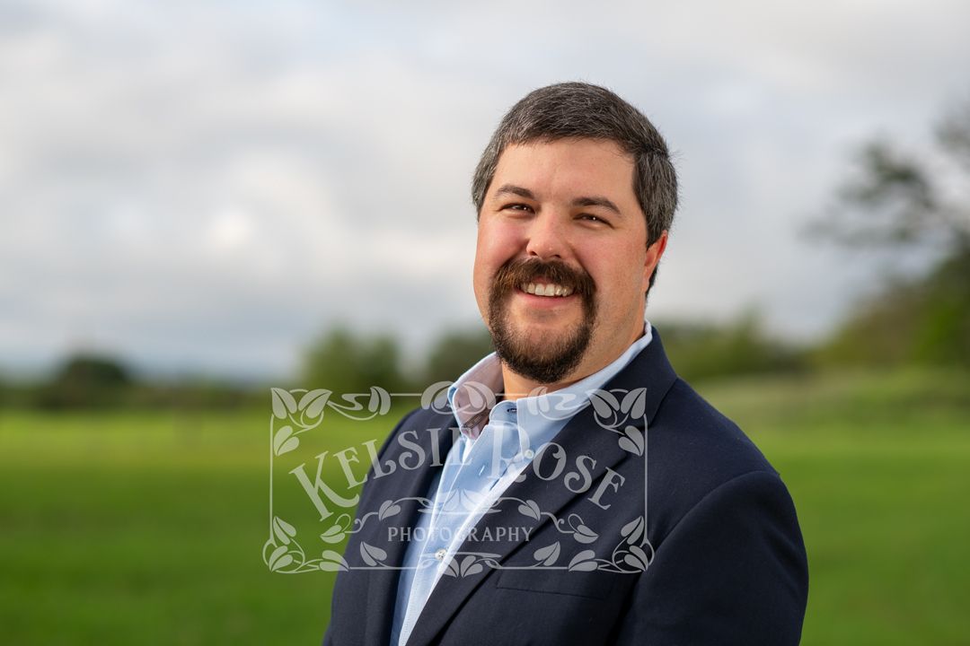 Headshot of smiling bearded businessman in navy suit posing outdoors in green field for professional portrait.