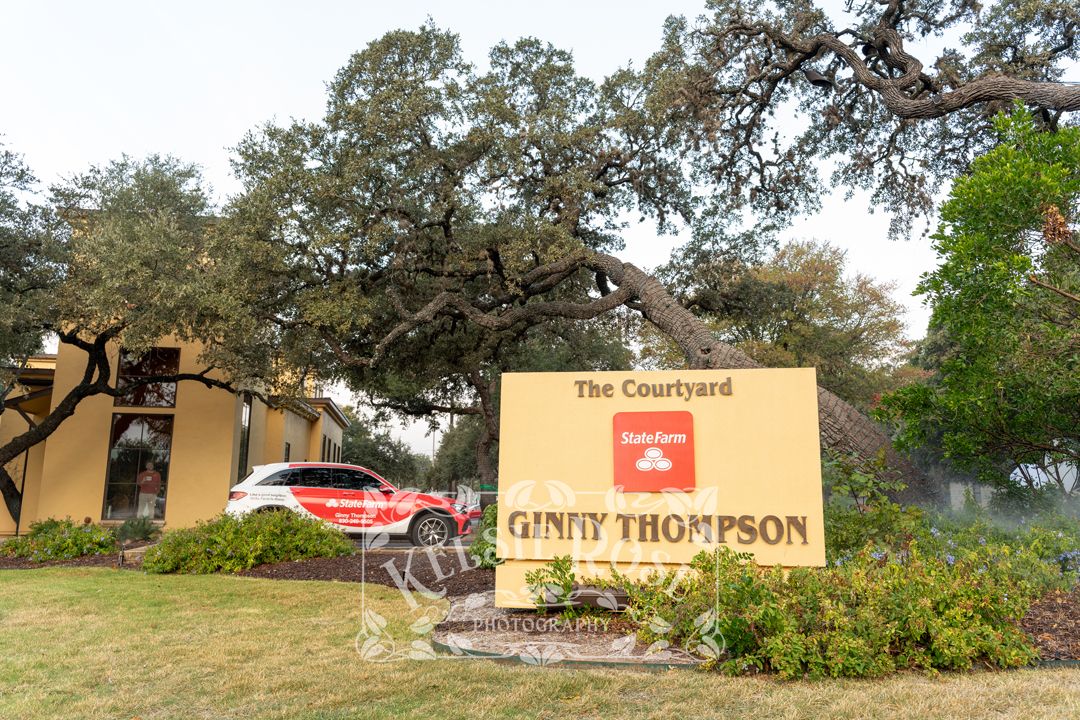 Sign for The Courtyard Ginny Thompson State Farm insurance office surrounded by large oak trees.