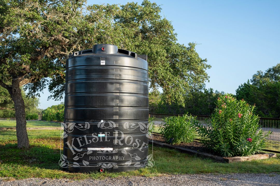 Black water storage tank, surrounded by trees and oleander bushes in a rural Texas setting.