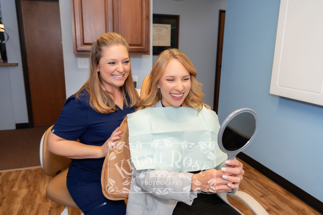 Dental hygienist assisting a smiling blonde patient reviewing her results in a handheld mirror.
