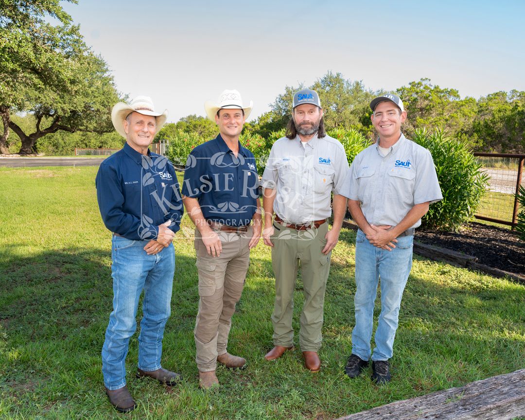 Group of five employees in uniform pose outdoors with grass and oak trees in the background on a sunny day in a rural Texas setting.