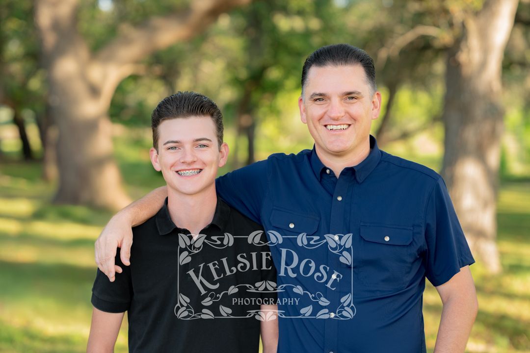 Father and son smiling and posing together in a sunlit green outdoor setting.
