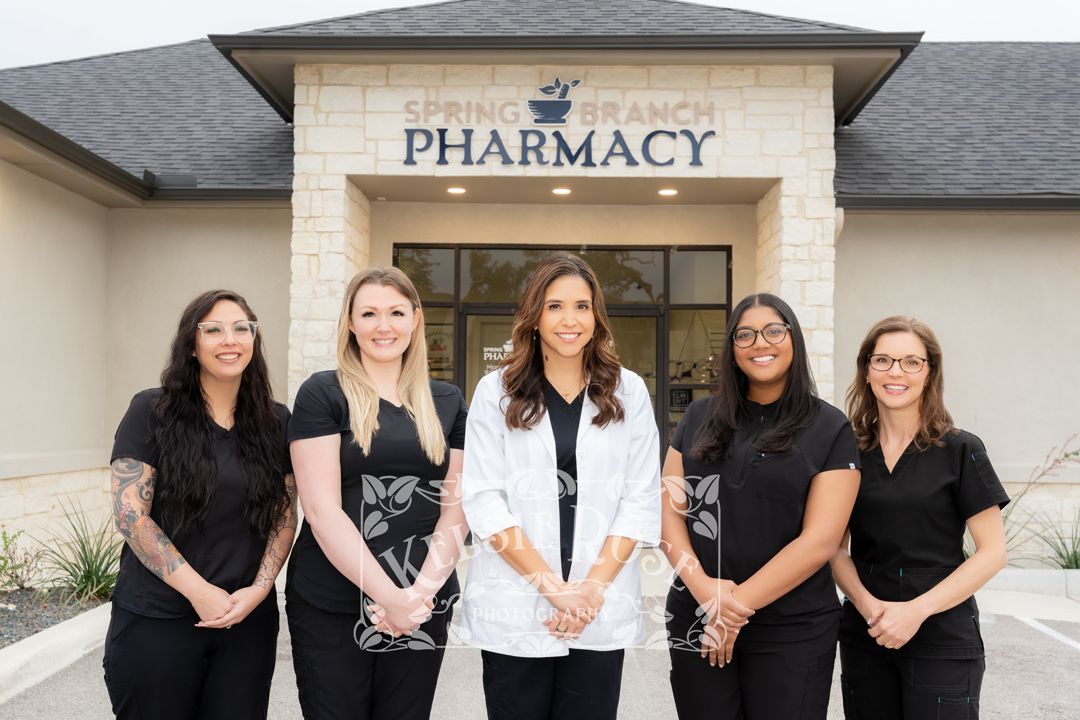 Five pharmacy staff members smiling in front of a pharmacy building, with one woman in a white coat centered among four in black scrubs.