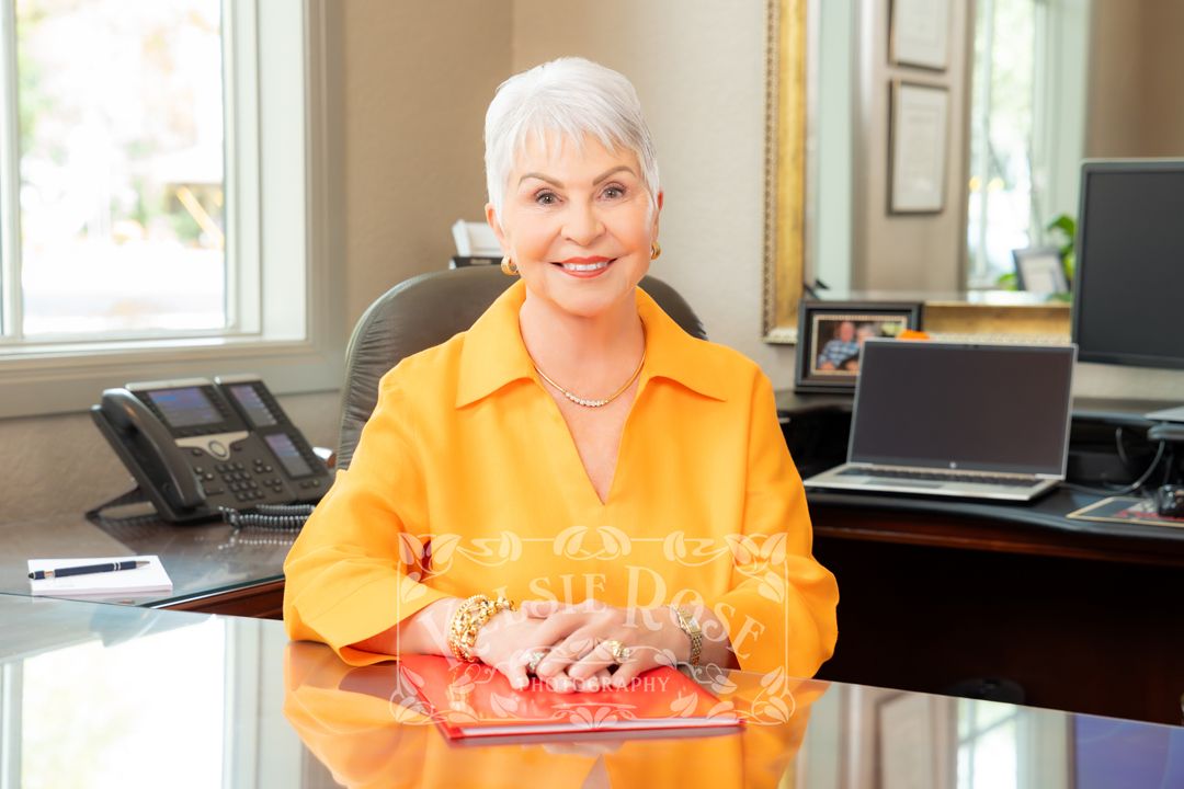 Professional woman in yellow blazer smiling at glass desk in modern office setting.