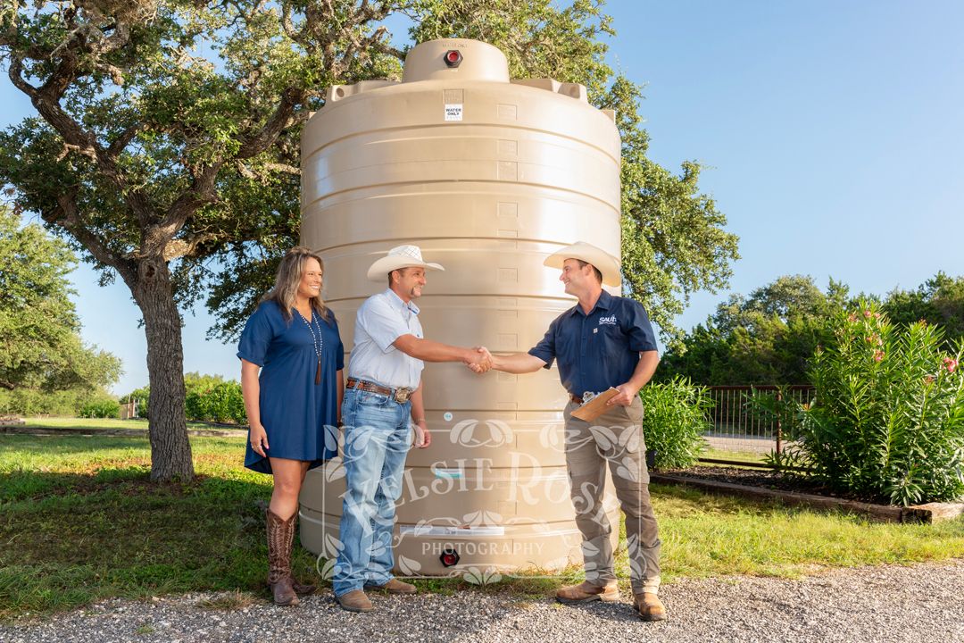Two people shaking hands near a large tan water storage tank outdoors, with a woman in blue dress standing beside them.