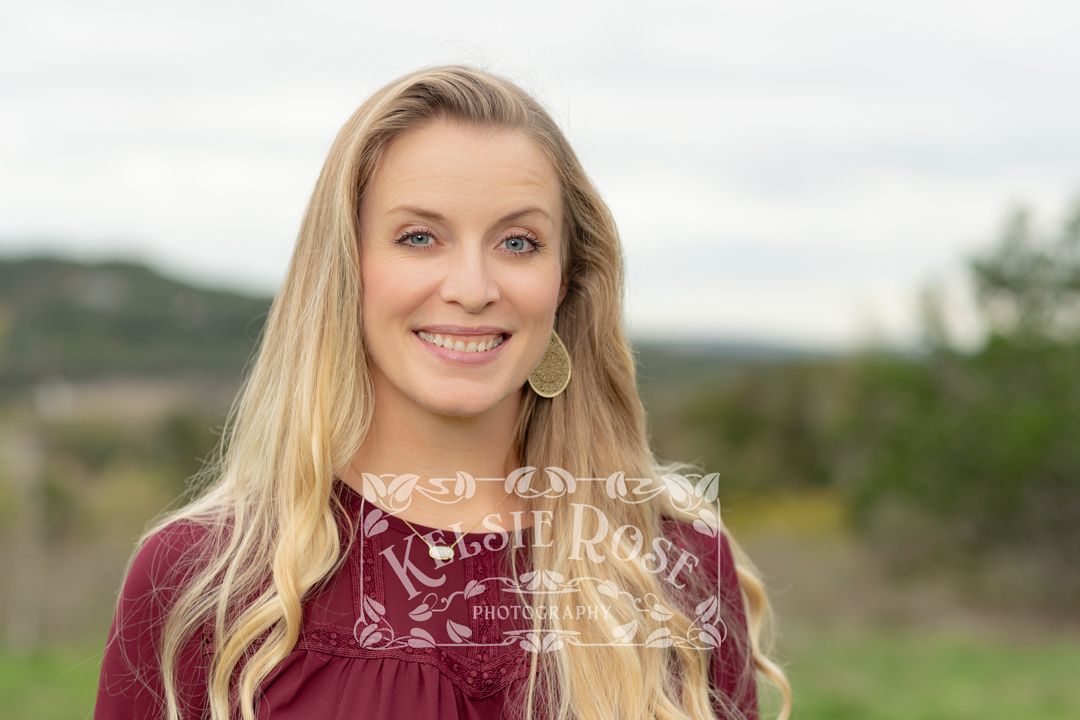 Headshot of smiling blonde woman in burgundy top posing outdoors with scenic Texas hill country behind her.