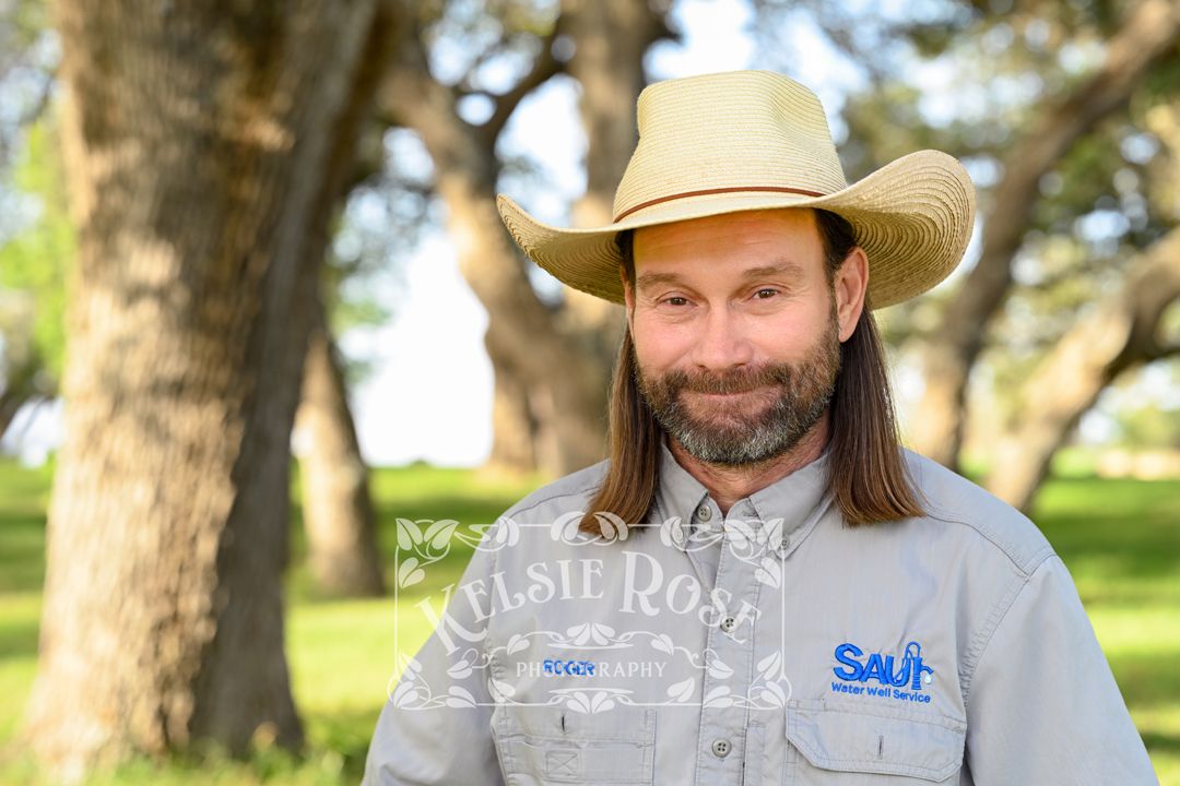 Headshot of bearded man with long hair wearing straw cowboy hat and water well company work shirt outdoors.