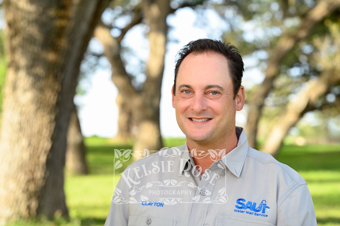Headshot of smiling man in water well company uniform shirt standing outdoors among trees.