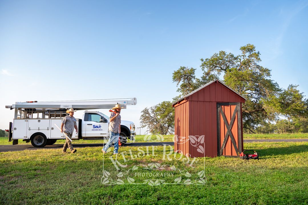 Two workers walking and carrying ladder in front of Saur Water Well Service truck and red well building on ranch.