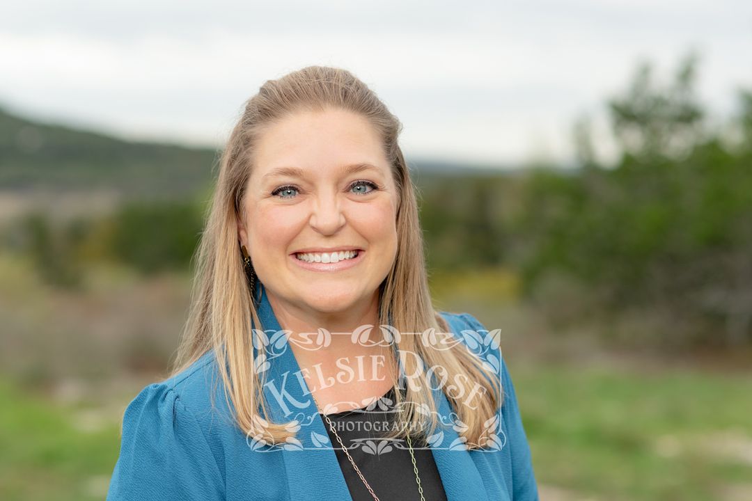 Headshot of smiling blonde woman in teal blazer smiling outdoors with Texas hill country and cloudy sky in background.