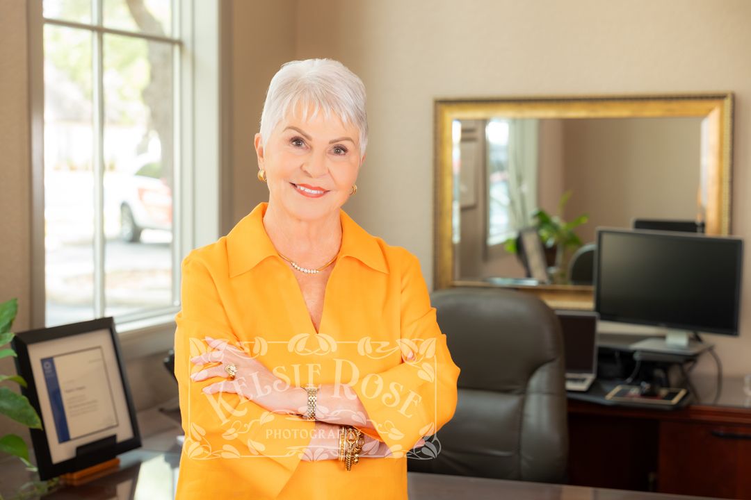 Older woman with white hair in yellow jacket stands arms crossed smiling in professional office setting.