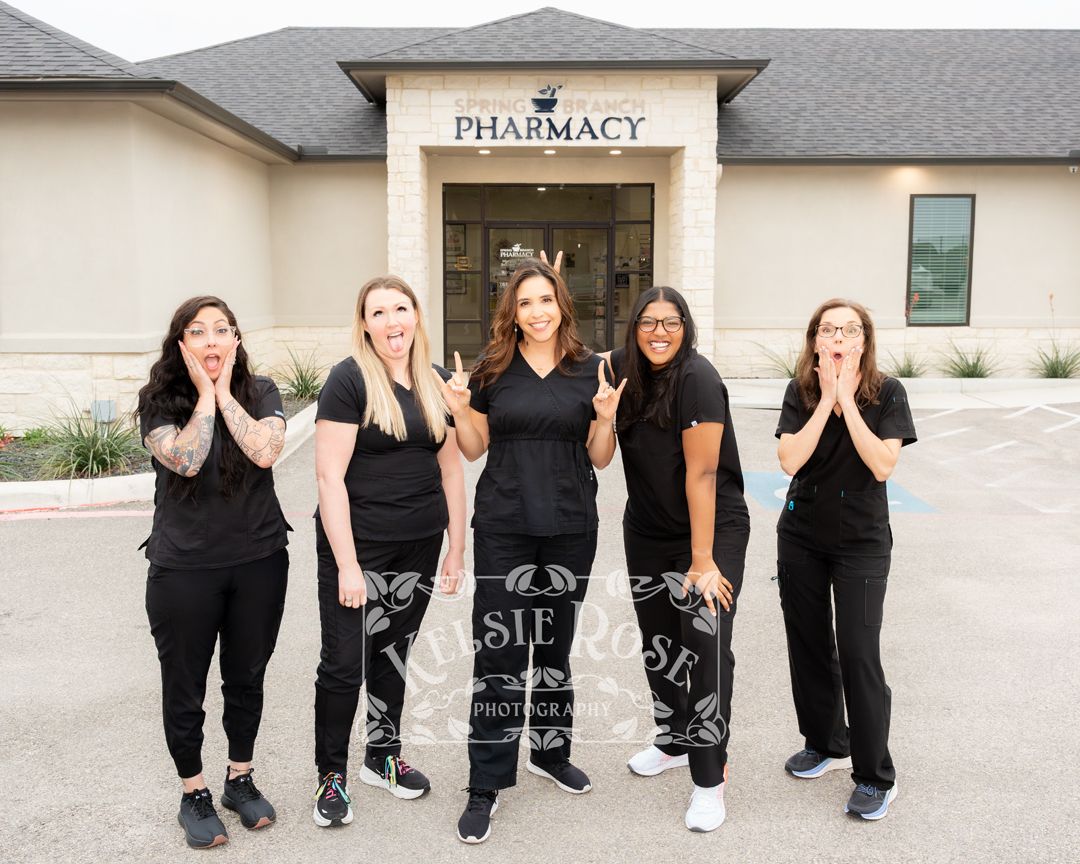 Five pharmacy staff in black uniforms pose playfully outside a pharmacy building, making surprised and silly expressions.