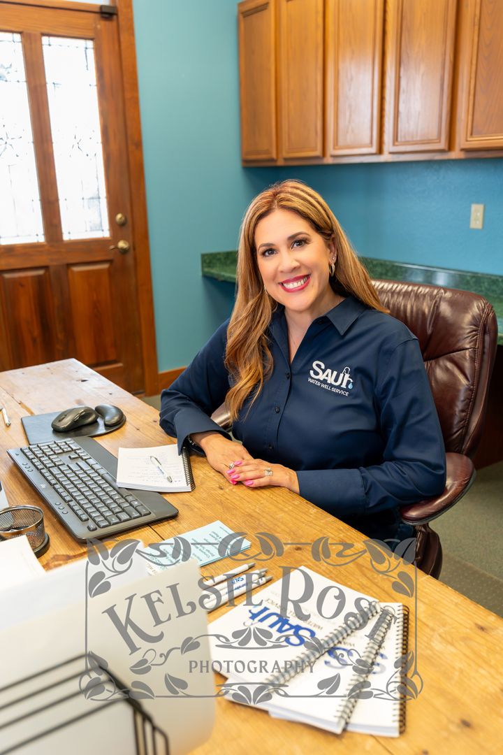 Smiling woman in Saul Water Well Service uniform sitting at office desk with computer and paperwork.
